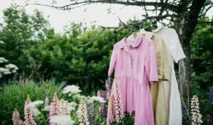 Natural Colored Dresses Hanging On On A Tree In The Garden With Lupine Flowers. Concept Organic Clothes